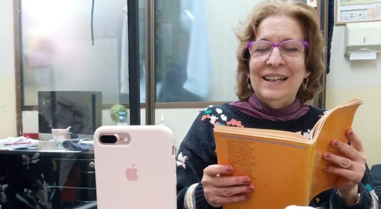 Una mujer con un libro al frente de su celular para grabar un audio leyéndolo