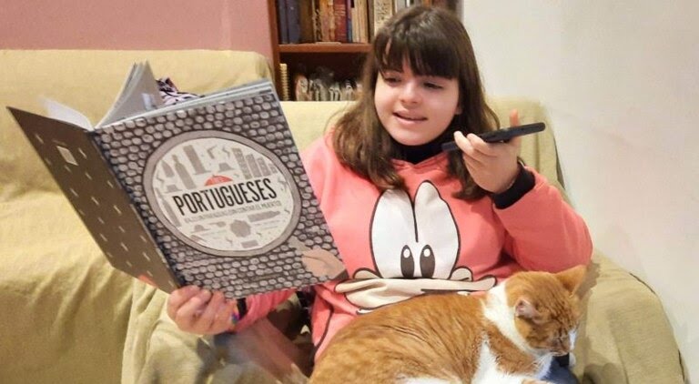Una niña con un libro y un celular grabando un audio leyendo el libro