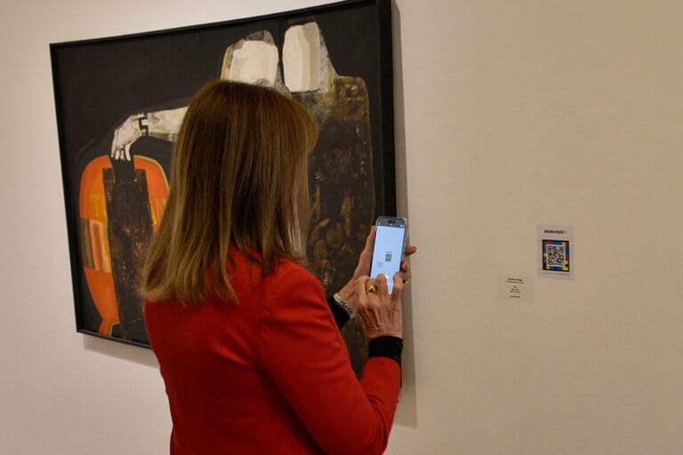 La imagen muestra a una mujer con su celular apuntando al código QR para saber la información que hay sobre el cuadro a su izquierda. Fuente: Fotografía de la página web del museo Emilio Caraffa
