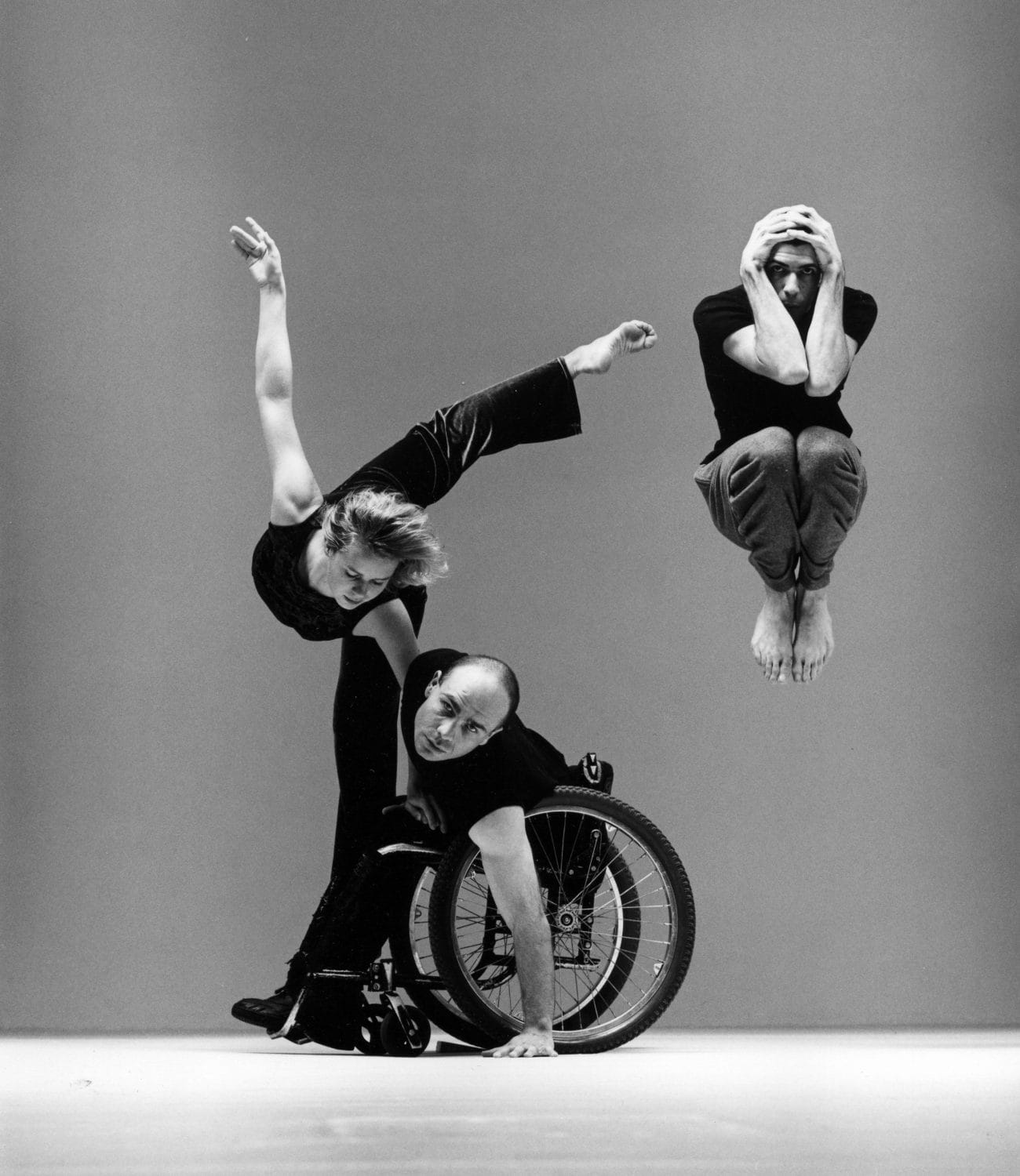 La imagen muestra a tres bailarines: en el medio a un hombre en silla de ruedas, a la izquierda una chica levantando una pierna y una mano y a la derecha, un hombre haciendo un salto agarrándose la cabeza. Fuente: Candoco Dance Company.
