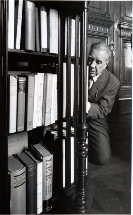 Fotografía en blanco y negro de Jorge Luis Borges en la Biblioteca Nacional. Está arrodillado al costado de un estante de libros.