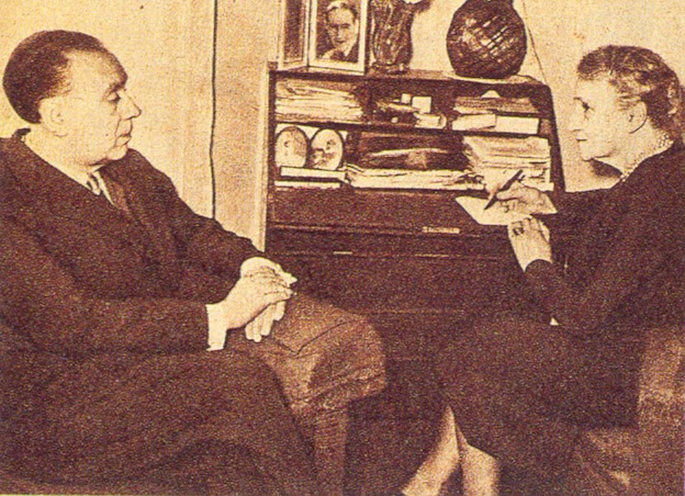Fotografía de Jorge Luis Borges sentado en un sillón, a la izquierda, dictándole a una mujer que está sentada a la derecha.
