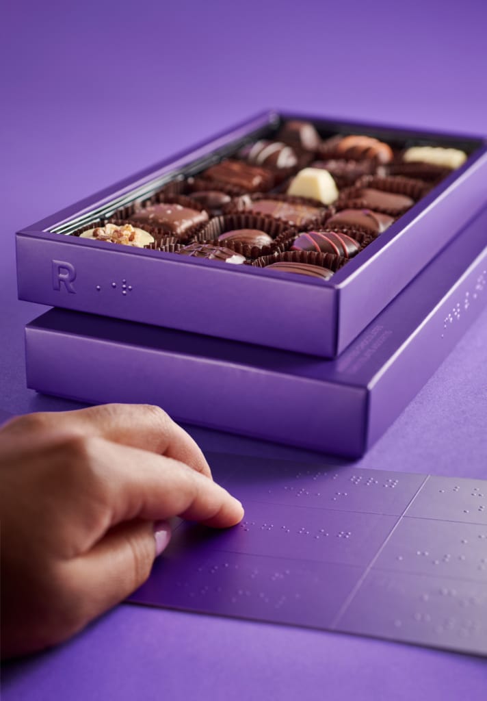 Se muestra una caja de bombones violeta totalmente accesible con pestañas de orientación en braille, una leyenda de bombones en braille y un código QR que enlaza con un lector de pantalla con una leyenda de audio.